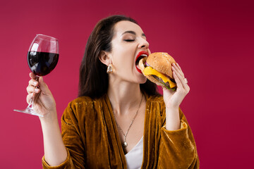 brunette woman in velour dress holding glass of wine and eating tasty burger isolated on red.