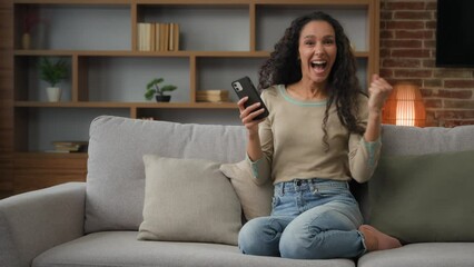 Happy cheerful woman sitting at home couch win with phone cellular device gadget betting good news victory mobile online winning screaming with shock yes gesture celebrate achievement with smartphone