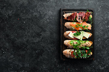 sandwich Set bruschetta with tuna, cheese, prosciutto and tomatoes on a dark wooden board. On a black stone background.