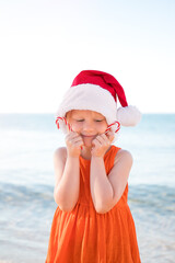 Girl in an orange dress, wearing a Santa Claus hat and holding Christmas lollipops on the beach is happy. Merry Christmas and Happy New Year.