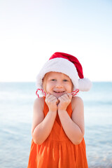 Obraz premium Girl in an orange dress, wearing a Santa Claus hat and holding Christmas lollipops on the beach is happy. Merry Christmas and Happy New Year.