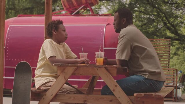 Medium slowmo of Black man with teenage son sitting at wooden street food cafe table outdoors in park drinking orange juice and chatting, spending weekends together