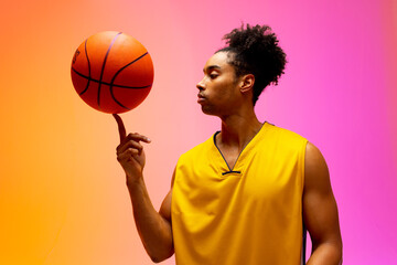 Image of biracial basketball player spinning basketball on pink to orange background - Powered by Adobe