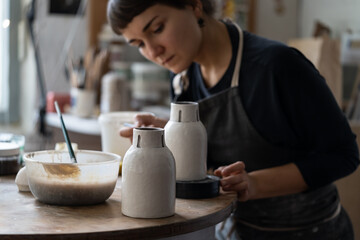 Young female artisan in black apron works with crockery sitting at round table in workshop against bright window. Brunette woman enjoys painting handmade ceramic vase in pottery craft studio