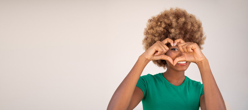 beautiful smiling dark-skinned woman in a green t-shirt playfully and flirting shows heart shape with hands. Happy positive love romantic emotions. afro hair style. Light beige background, copy space.