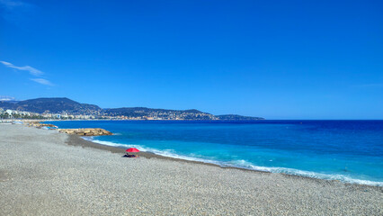 Calme ou solitude ? Seulement un moment de quiétude sous un parasol rouge sur la plage de galets de Nice sur la Côte d'Azur face aux couleurs bleues de la mer Méditerranée 