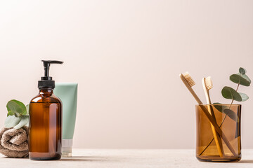 Bamboo toothbrushes in glass, tube of toothpaste and liquid soap in glass jar on light background....