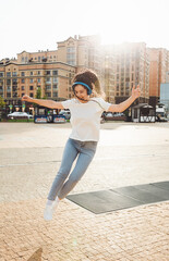 A cheerful woman listens to music through headphones outside on a sunny day. a girl with headphones jumps on the street. generation z