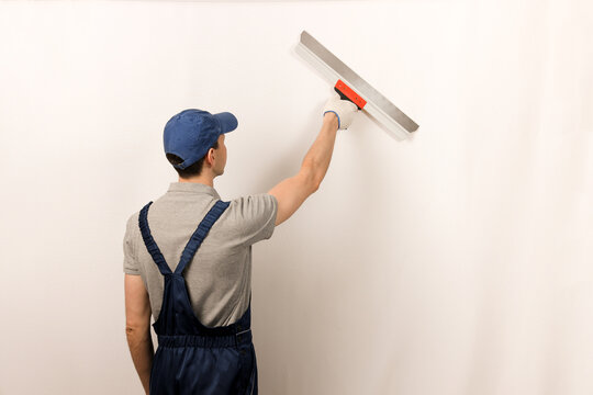 Worker in wearing overalls and cap plastering a wall with putty using a putty knife. Plasterer in blue working uniform plastering the wall indoors. Repair work and construction concept.