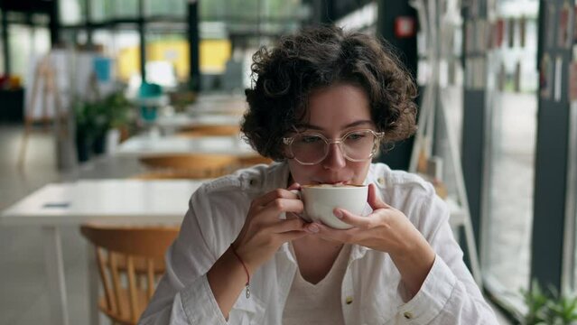 Young bright woman drinks coffee sits in a restaurant holding a cup of cappuccino with two hands Lunch break drinks coffee in a bright cafe Portrait of a young girl who is having lunch on a bright day