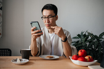 Adult asian man wearing glasses looking at smartphone during breakfast. Leisure morning time.