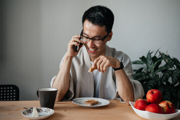 Fototapeta na wymiar Smiling adult asian man wearing glasses talking on smartphone during breakfast. Leisure morning time.