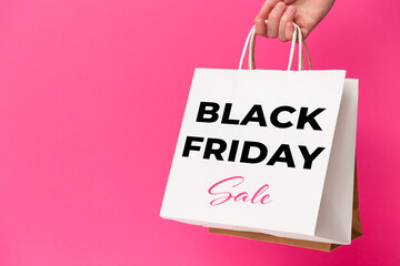 Female hand holding white shopping paper bags with Black Friday Sale text on pink background. Sale,...