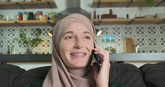 Muslim woman with braces communicates on the phone in the living room at home. Portrait, girl smiling, happy. The concept of communication with loved ones, family, husband.