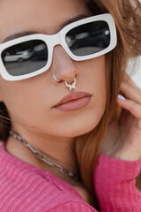 Naklejka premium Fresh summer fashion close-up portrait of a stylish beautiful young woman with trendy white sunglasses with a nose piercing wearing a pink knit top. Clear female face