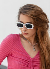 Naklejka premium Cool trendy pretty redhead girl with white fashion sunglasses in a stylish pink knit top walking outside on a sunny summer day
