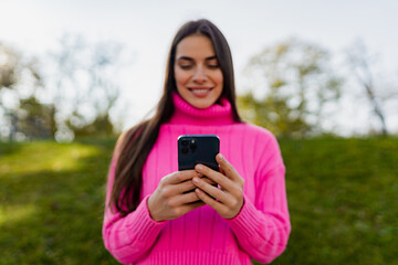 young smiling woman in pink sweater walking in green park using phone