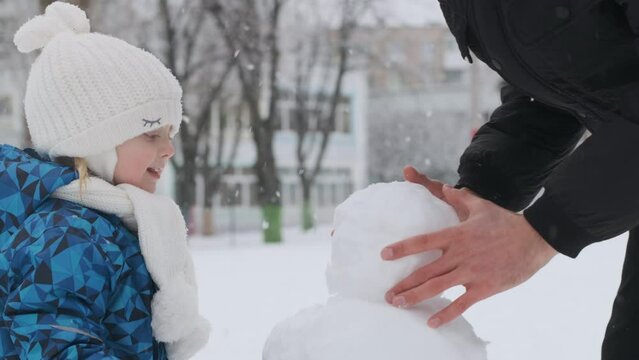 Little girl and man make snowman outside during snowfall. Dad and daughter play outside in the winter.