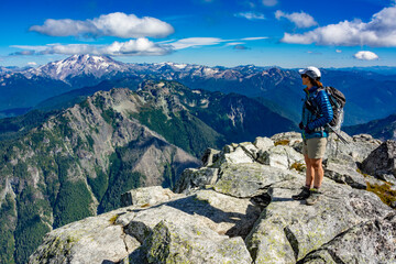 Fototapeta premium Adventurous athletic female hiker standing on top of a rugged mountain in the Pacific Northwest with jagged mountains in the background. 