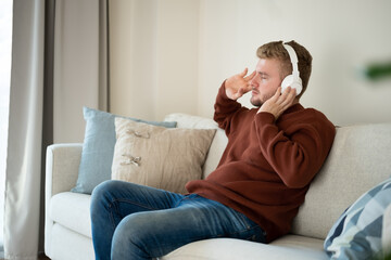 Fototapeta premium Young caucasian blond curly hair man listening to music with big white handsfree headphones sitting on sofa at home on neutral white background. Handsome adult guy portrait lifestyle.Copyspace.