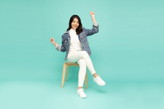 Attractive Beautiful Asian Woman Sitting On White Modern Chair And Hands Up Raised Arms From Happiness, Full Body Composition