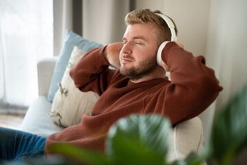 Young caucasian blond curly hair man listening to music with big white handsfree headphones sitting...
