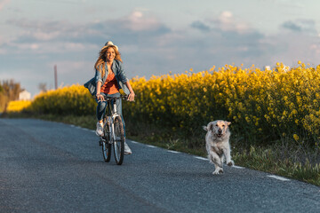 Shot of beautiful young girl riding a bike while walking her dog in the park through rapessed field.