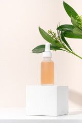 Mockup cosmetics serum with a pipette in a transparent frosted glass and orange liquid. Facial peeling with lactic Aha acid on a white cubic pedestal, beige background with leaves