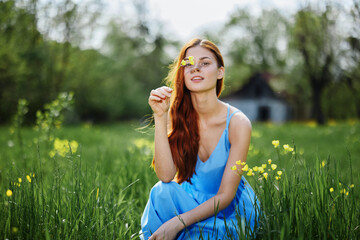 Naklejka na ściany i meble Woman in spring in nature smile with teeth sitting in a field on the green grass with flowers and smiling happy sunset in a blue dress with red hair
