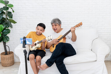 Asian family, father and son are sitting on the sofa is playing guitar through online channels by...