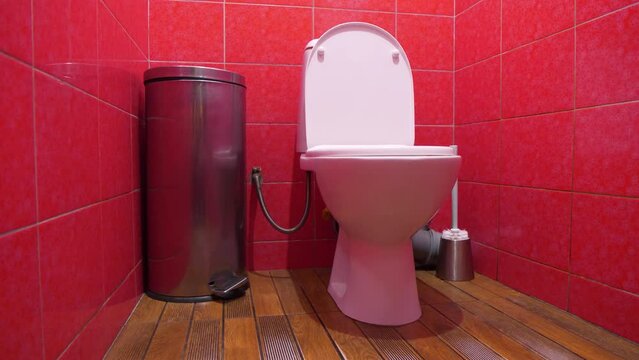 Toilet room interior with white toilet bowl with open lid, metallic wastebasket, toilet brush and red wall tiles