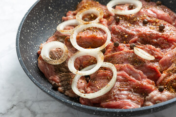 Raw beef with onions marinated with spices in a frying pan on a light texture background. Meat recipe. Juicy grilled steak, close-up. Place for text, space for copy.
