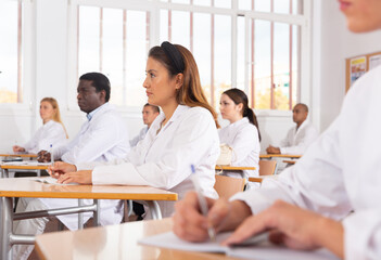 Positive latin american girl in white coat listening to lecture and taking notes in classroom...