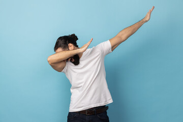 Portrait of anonymous unknown man with beard wearing white T-shirt standing in dab dance pose, internet meme, celebrating success. Indoor studio shot isolated on blue background.