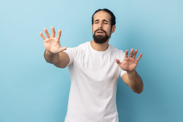 Portrait of disoriented blind man with beard wearing white T-shirt standing with stretched arms, searching for way, vision problems. Indoor studio shot isolated on blue background.