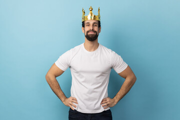 Portrait of confident man with beard wearing white T-shirt and gold crown, standing with hands on...