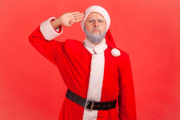 Fototapeta na wymiar Yes, sir! Portrait of serious obedient elderly man wearing santa claus costume saluting with arm near temple and listening attentively to order. Indoor studio shot isolated on red background.