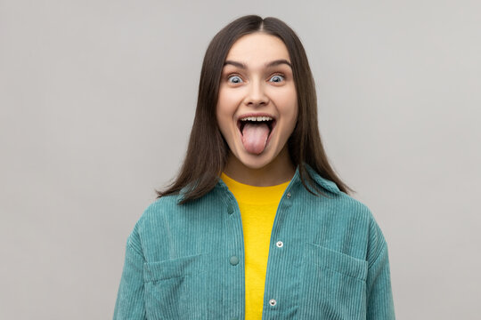 Dark haired woman sticking out tongue and looking at camera, teasing with naughty expression, disobedient behavior, wearing casual style jacket. Indoor studio shot isolated on gray background.