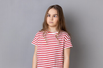 Portrait of funny silly little girl wearing striped T-shirt looking up cross eyed with stupid dumb face, girl has awkward confused comical expression. Indoor studio shot isolated on gray background. - obrazy, fototapety, plakaty
