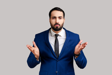 Portrait of bearded man standing with raised hands and confused indignant expression, asking why how, what reason, wearing official style suit. Indoor studio shot isolated on gray background.