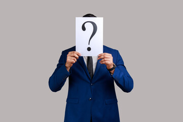 Unknown man hiding his face behind white paper with question mark, finding smart solution, asking for advice, wearing official style suit. Indoor studio shot isolated on gray background.