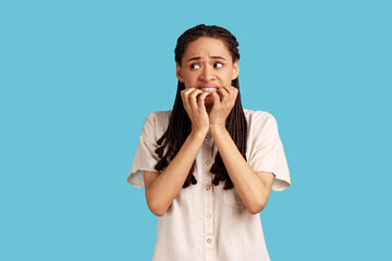 Portrait of puzzled woman with black dreadlocks has worried nervous expression, keeps hand on mouth, hears unexpected bad news, wearing white shirt. Indoor studio shot isolated on blue background.
