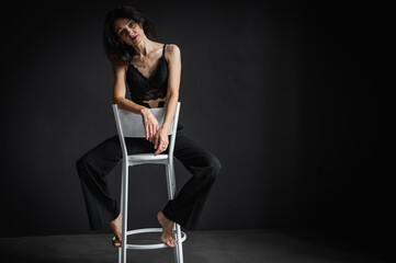 Full body photo of cute and seductive brunette woman sitting in pants on chair isolated on dark background 