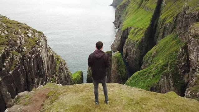 4K Aerial drone view of a man looking down to Dunnesdrangar, Faroe Islands. Amazing nature, huge cliffs and mountains in Faroe Islands. Vagar Island. Many flying sea birds. Sunny day in summer.