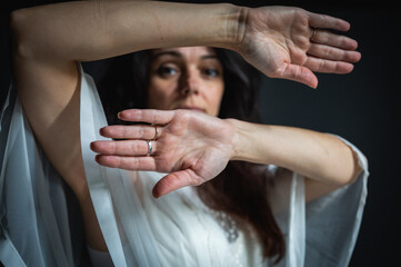 Anxiety, danger, protection - conceptual image of a woman covering her face with her hands, looking through them and peeking with one eye between her fingers on a dark studio background