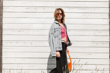 Stylish beautiful young woman with orange sunglasses in a fashionable denim jacket and bag skirt walks outdoors near a white vintage wooden wall