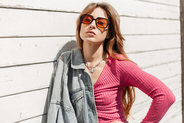 Fashion fresh sunny portrait of a beautiful stylish young girl with cool sunglasses in a fashionable denim jacket and pink top stands near a white village timber wall outdoors