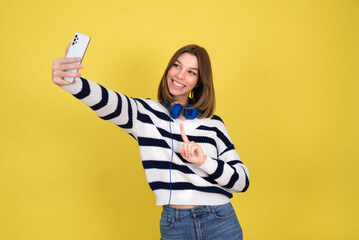 Funny teenage  girl making selfie. Smiling girl wearing  holding  smartphone, making faces on camera, posing for selfie . yellow  background