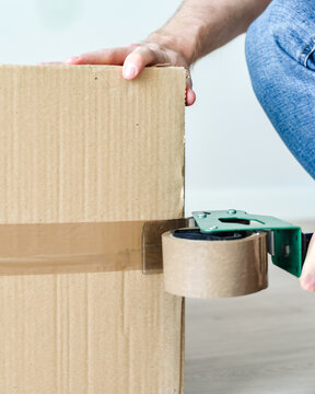 Detalle de un hombre usando una herramienta para precintar una caja de cart&oacute;n con cinta adhesiva marr&oacute;n. Embalaje de cajas de mudanza.