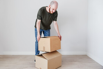 Un hombre joven con barba agarra una caja de cartón. Concepto de mudanza y amueblar un nuevo hogar. - obrazy, fototapety, plakaty